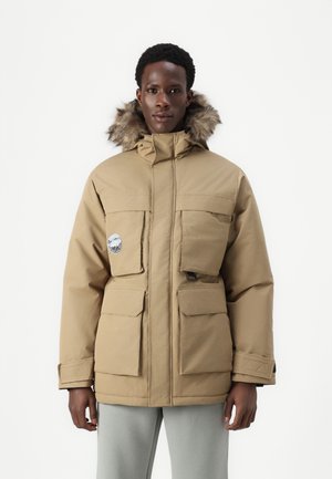 Beige Parkajacke mit gefütterter Kapuze aus Fell, verfügt über mehrere Vordertaschen und ein rundes Logopatch, gestaltet mit einem geraden Schnitt und gerippten Bündchen.