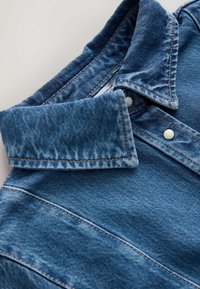 Gros plan sur le col d'une veste en denim bleu pliée, avec des coutures visibles et des boutons-pression en métal sur un fond clair.