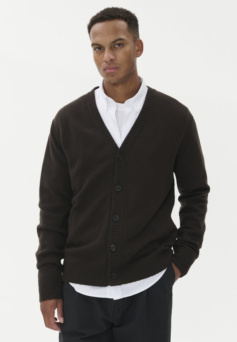 Brūns adīts kardigans ar V-veida kaklu, ribotām piedurknēm un sešām pogām. Valkāts pār baltu krekli, attēlots uz vienkrāsaina balta fona.