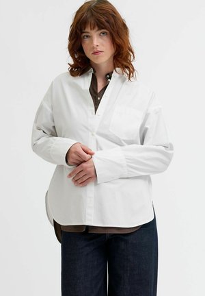 Weißes Langarmhemd aus Baumwolle mit Knopfleiste, einer Brusttasche und einem lässigen Schnitt, getragen über einem braunen Hemd und dunklen Jeans.