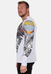 Weißes Langarmhemd mit einem großen Adlerdesign und Text auf den Ärmeln. Das Design umfasst sechseckige Muster und kräftige Farben.