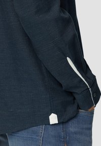 Chemise à manches longues bleu foncé texturée avec des accents blancs au niveau des poignets. Présente un bouton de fermeture et un détail d'ourlet distinct. Portée avec un jean bleu.