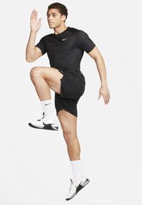 Pantalones cortos deportivos negros y camiseta, calcetines blancos y zapatillas negras y blancas. La persona está en una pose dinámica de carrera, levantando una rodilla.