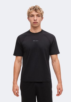 Ung mand med kort blondt hår iført en sort Champion t-shirt og sorte bukser står mod en ensfarvet lys baggrund.