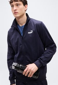 Μπλε σκούρο μπουφάν PUMA με φερμουάρ, ψηλό γιακά, στενή εφαρμογή και λευκό λογότυπο. Μαύρο παγούρι νερού με διαφανές καπάκι κρατημένο στο χέρι.