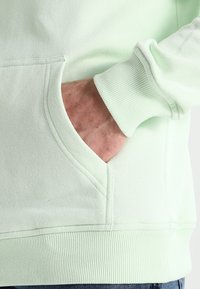 Una mano parzialmente all'interno della tasca anteriore di un felpa verde chiaro con polsini e orlo a coste, su uno sfondo semplice.