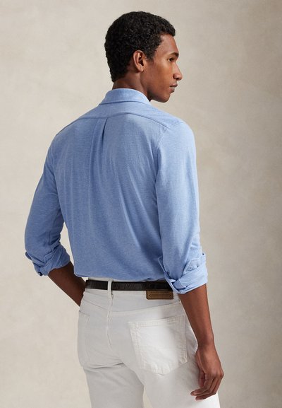 Άνδρας που φοράει ανοιχτό μπλε πουκάμισο με μακριά μανίκια σηκωμένα και λευκό παντελόνι, στέκεται με την πλάτη και το προφίλ στο πλάι να είναι ορατά.