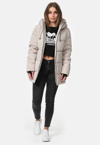 Doudoune beige clair avec capuche ajustée, fermeture éclair à l'avant et poignets élastiques, portée par-dessus un sweat-shirt noir court et un jean noir.