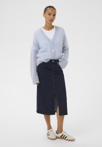 Hellblauer, flauschiger Cardigan mit Knöpfen, kombiniert mit einem dunkelblauen Jeansrock mit Frontschlitz und weißen Sneakern mit schwarzen Streifen.