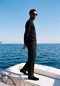 Homme portant des vêtements noirs et des lunettes de soleil debout sur le pont d'un bateau avec l'océan et un ciel clair en arrière-plan.