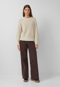 Frau steht vor einem einfarbigen Hintergrund und trägt einen cremigen Strickpullover, weite braune Hosen und schwarze Stiefel mit hellbraunen Sohlen.