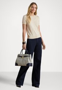 Béžový pletený sveter s krátkymi rukávmi, kombinovaný s tmavomodrými širokými nohavicami. Drží textúrovanú sivú kabelku s tmavomodrými doplnkami a zlatou kovovou výbavou.