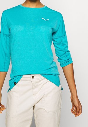 Osoba ubrana w turkusową koszulę z długim rękawem z małym białym logo na piersi oraz jasnobeżowe spodnie z przednimi kieszeniami.