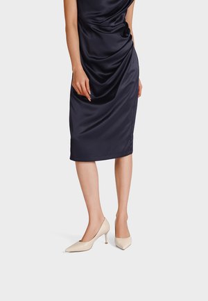 Donna che indossa un vestito midi senza maniche in raso blu navy e décolleté crema con tacco kitten a punta, in piedi davanti a uno sfondo neutro.