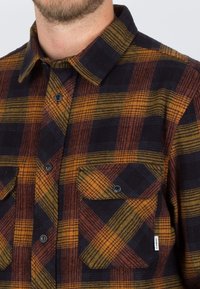 Camisa de franela con estampado a cuadros en azul oscuro, naranja y amarillo. Cuenta con dos bolsillos en el pecho y botones negros. Textura suave.