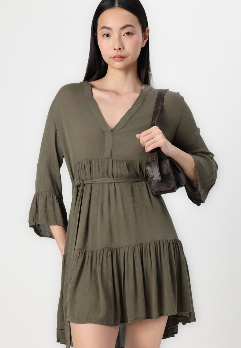 Robe vert olive avec un décolleté en V, un ourlet à volants en étages et des manches cloche trois-quarts, accompagnée d'un petit sac à main marron.