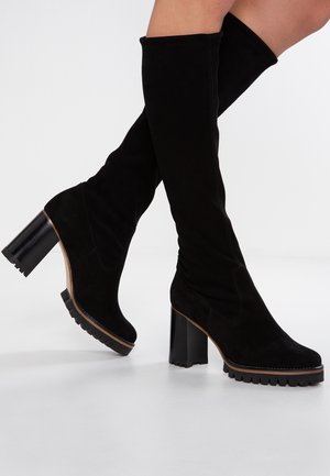 Bottes en daim noir montant jusqu'au genou, avec talons épais et semelles à crampons, portées sur des jambes croisées sur fond blanc uni.