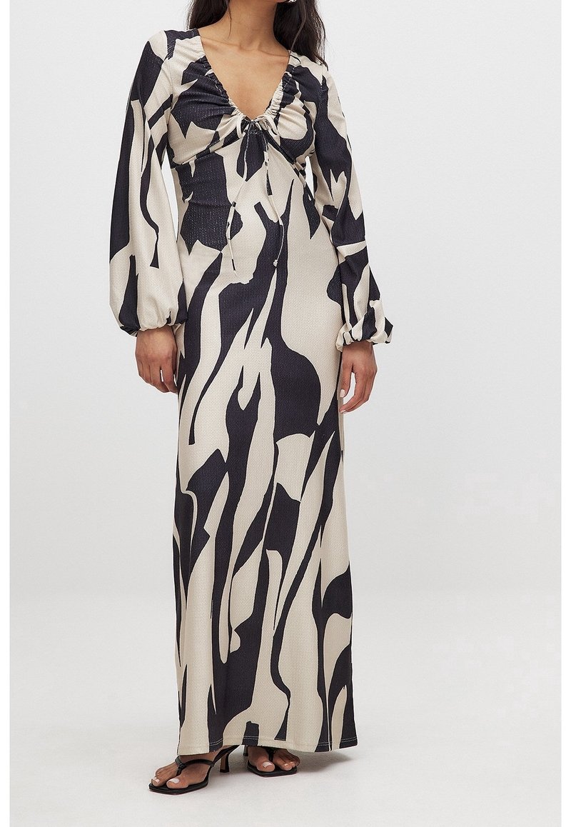 Femme portant une robe longue à manches longues, à imprimé abstrait noir et blanc, col froncé et sandales à talons noires.