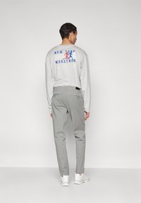 Lichtgrijs langarm shirt met de tekst "NEW YORK MARATHON" en een graphic van hardlopers op de achterkant, gecombineerd met lichtgrijze broek en witte sneakers.