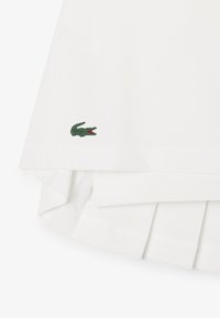 Falda blanca con pliegues hecha de tela suave. Presenta un logo de cocodrilo verde cerca del dobladillo. El dobladillo está limpio y terminado sin adornos.