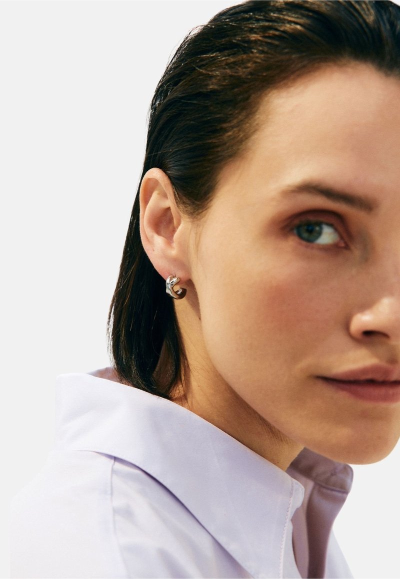 Hopeiset ja ruskeat nappikorvakorut, joissa on mutkikas muotoilu, yhdistäen sileän metallin kiiltävään ruskeaan yksityiskohtaan, esitettynä vaaleaa taustaa vasten.