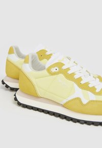 Gula sneakers med ovandel i mocka och mesh, vita snören, vadderad krage och texturerad svart gummisula. Logdetalj på sidan.