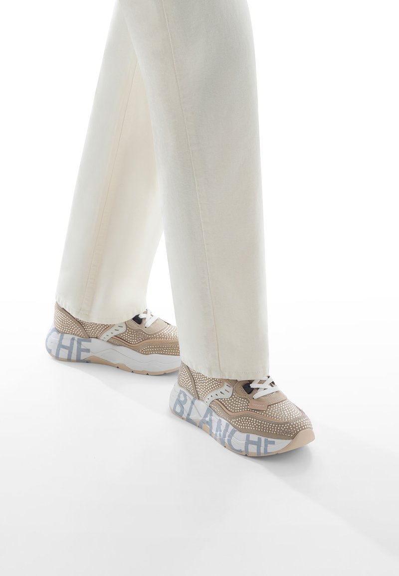 Beige Sneakers mit Netzdesign, weißen Schnürsenkeln und einer weißen Sohle mit "BLANCHE"-Text auf der Außensohle. Begleitet von cremefarbenen Hosen.