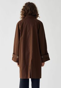 Bruine, lichte trenchcoat met een brede kraag en opgerolde mouwen. Heeft grote knopen en een achteropening voor extra bewegingsvrijheid.