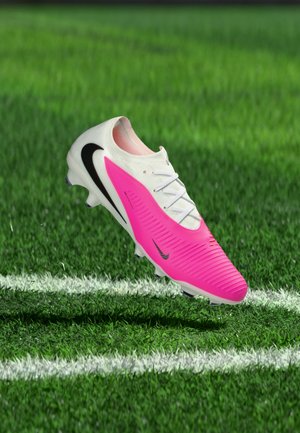 Chaussure de football Nike blanche et rose vif avec un swoosh noir, suspendue au-dessus de l'herbe verte près de la ligne blanche du terrain.