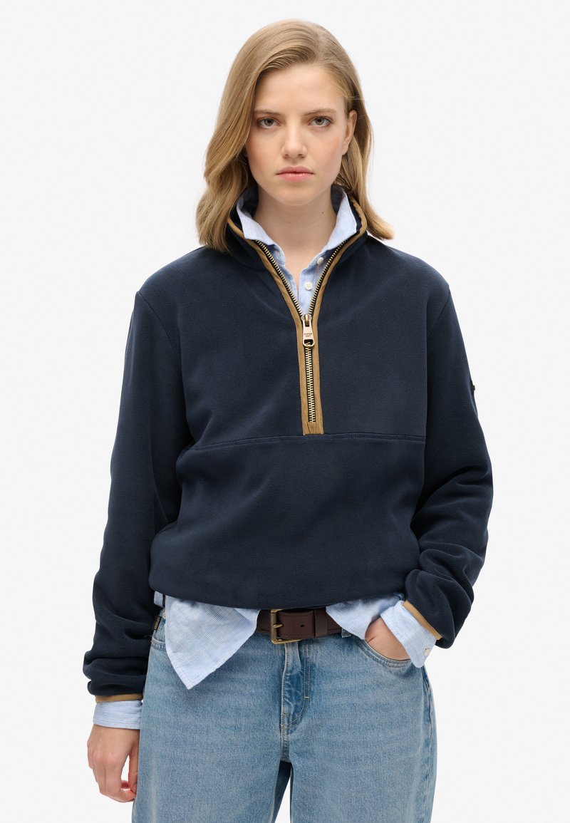 Marineblauwe fleece pullover met een halve ritssluiting en een beige rits, gedragen over een lichtblauw overhemd met knopen, gecombineerd met lichte spijkerbroeken.