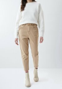 Suéter de punto suave color crema con hombros caídos, combinado con pantalones de tiro alto en tono beige y botas de tobillo beige. Los pantalones tienen bolsillos frontales.