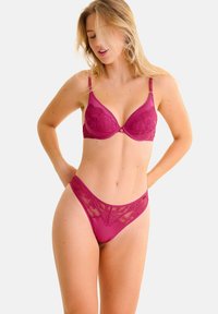 Magenta kant-bh met voorgevormde cups, verstelbare bandjes en decoratieve hardware aan de voorkant; bijpassende doorzichtige kant-string met bloemmotief.