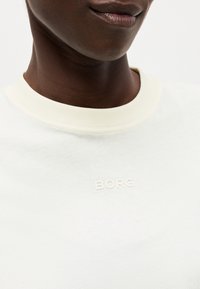 Κοντινό ενός ατόμου που φορά λευκό μπλουζάκι με στρογγυλή λαιμόκοψη και μικρό ανάγλυφο κείμενο "BORG" στο κέντρο του στήθους.