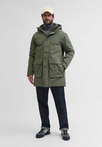 Olivno zelena vodoodporna parka s kapuco, dvema velikima sprednjima žepoma in zadrgo, oblečena čez sivo pulover s turtle neck in temne kavbojke. Bež kapica in čevlji z svetlimi vezalkami.
