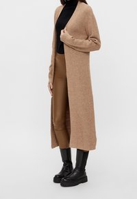 Langer, offen gestalteter Cardigan aus beigem Strick, mit strukturiertem Stoff, kombiniert mit braunen, schmal geschnittenen Hosen und schwarzen klobigen Stiefeln. Schlichtes, vielseitiges Design.