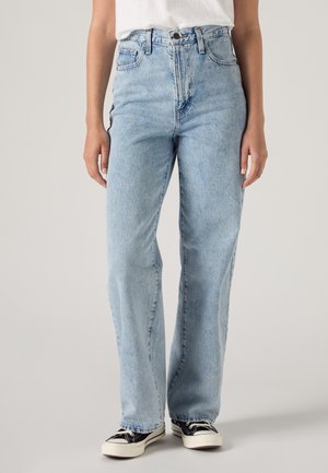 Un mannequin porte un jean délavé clair à coupe droite, un tee-shirt blanc et des baskets noires et blanches sur un fond de studio uni.