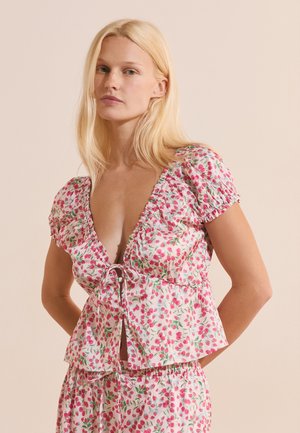 Femme blonde portant un haut blanc et un pantalon avec des motifs floraux roses, debout, les mains dans le dos, sur un fond uni.