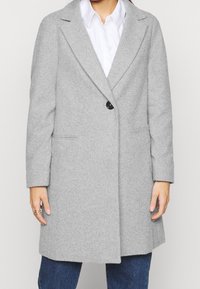 Cappotto grigio chiaro in misto lana con maniche lunghe, rever a punta, chiusura a un bottone e tasche laterali, abbinato a una camicia bianca.