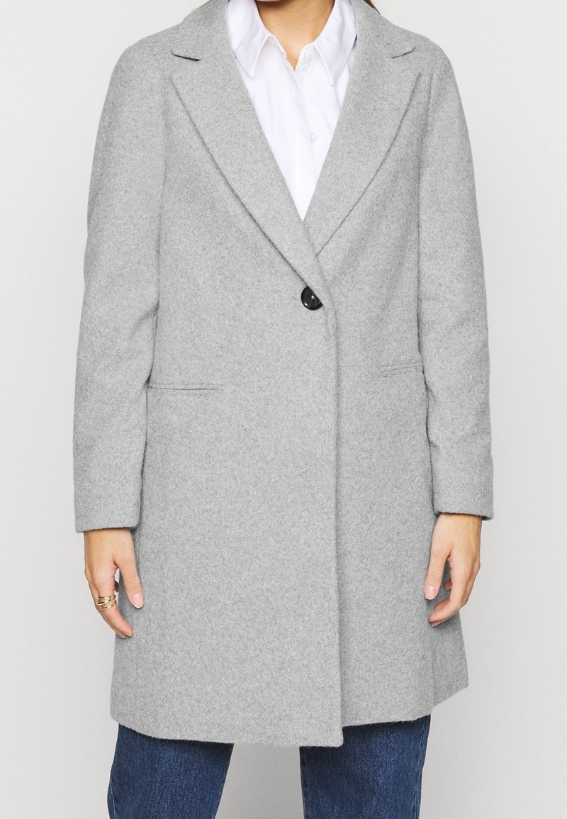 Cappotto grigio chiaro in misto lana con maniche lunghe, rever a punta, chiusura a un bottone e tasche laterali, abbinato a una camicia bianca.