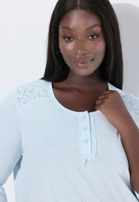 Žena s tmavou pokožkou a dlouhými tmavými vlasy, která má na sobě světle modrý top s krajkovými detaily na ramenou a knoflíky u výstřihu.