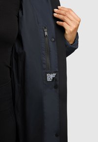 Tamsiai mėlynas švarkelis su užtrauktuku kišenėje, spaudėmis ir audinio etikete su užrašu „Khujo original for a few“. Lygi tekstūra su kontrastingomis detalėmis.