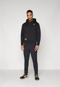 Sweatshirt preta com capuz, com logo, bolso frontal e cordões, combinada com calças pretas afuniladas e sapatilhas desportivas pretas e castanhas.
