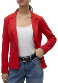 Blazer rouge ajusté avec revers, en tissu texturé, porté sur un haut blanc, associé à un jean bleu taille haute et une ceinture noire.