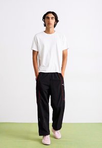 Witte t-shirt met korte mouwen, zwarte sportbroek met rode accenten en een logo, gecombineerd met lichtroze sneakers op een groene vloer.