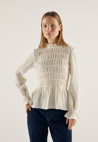 Witte smocked blouse met een gerimpelde hoge halslijn, lange mouwen en een geplooid textuur; gecombineerd met donkere jeans. Lichtgewicht, textuurstof.