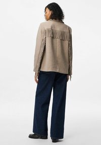 Beež villane jakk, millel on fringing detailid seljal ja varrukatel, mida on paaritatud tumehallide laiemate pükstega ja mustade säärebotastega.