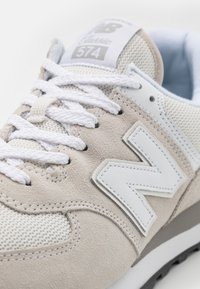 Beige ja valkoinen New Balance 574 -kenkä, jossa on mokkanahkaa, verkkokangasta, valkoiset nauhat ja sivussa brodeerattu "N"-logo.