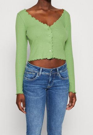 Person iført en lysegrøn ribbet, knaplukket crop top med lange ærmer og flæsekant samt blå denimjeans, der står mod en hvid baggrund.
