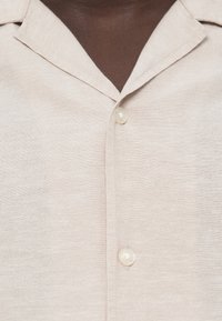 Φωτεινό μπεζ πουκάμισο με κοντό μανίκι, γιακά τύπου spread, κλείσιμο με κουμπιά μπροστά και υφή υφάσματος. Διαθέτει λείο, απαλό φινίρισμα.