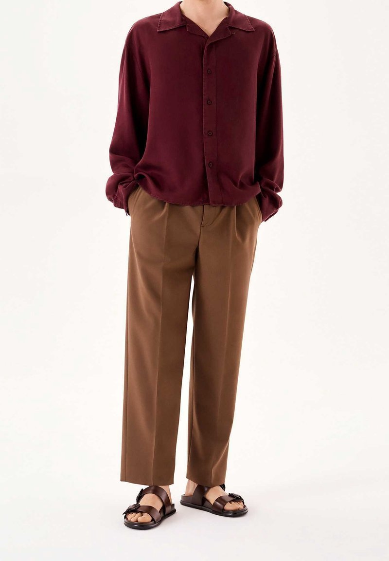 Personne portant une chemise bordeaux ample à boutons, un pantalon plissé marron et des sandales marron foncé, debout devant un fond blanc.